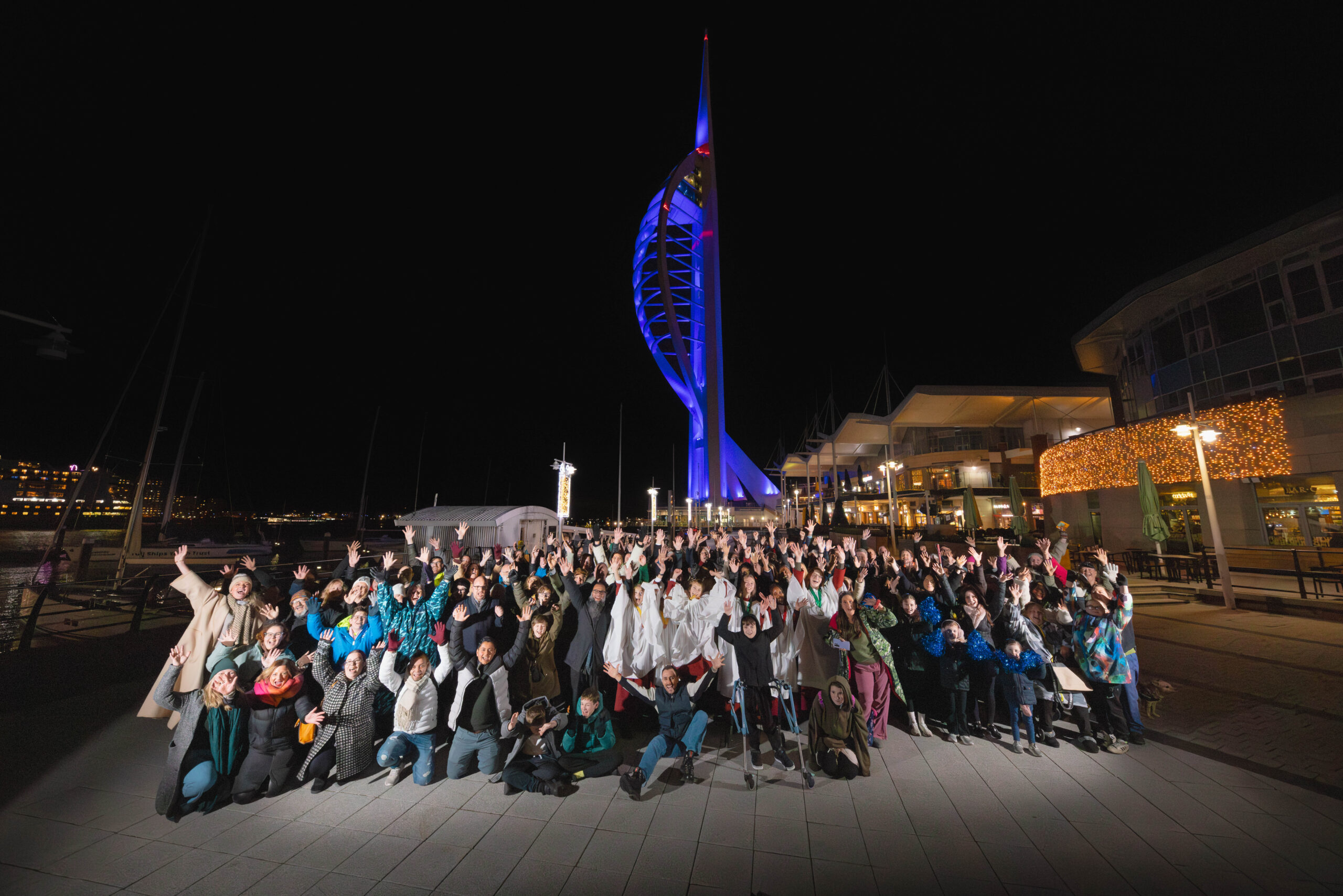 A large crowd of people, cheering with arms up in the air in front of a blue lit Spinnaker Tower.